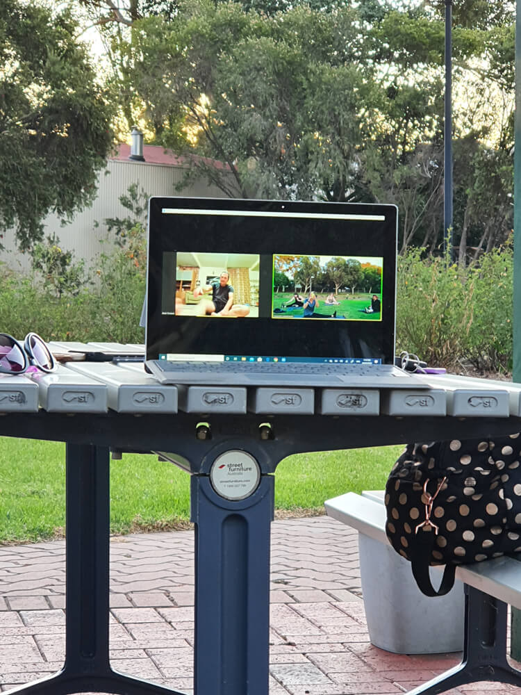A laptop on a table outside in a park with a fitness class streaming on the screen.