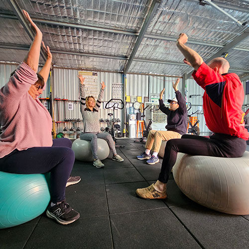 Trainer Cass and 3 NDIS clients sitting on fit balls inside a gym with arms stretched high.